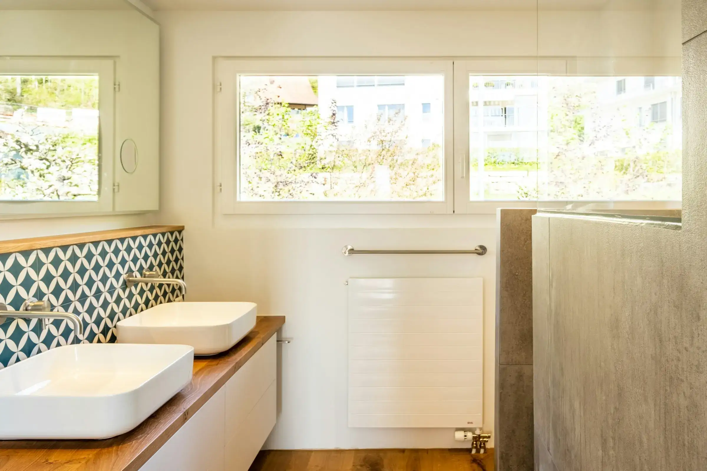 After: bright bathroom with a wide new window, two white basins on an oak counter, teal patterned tile and a concrete-look shower wall.