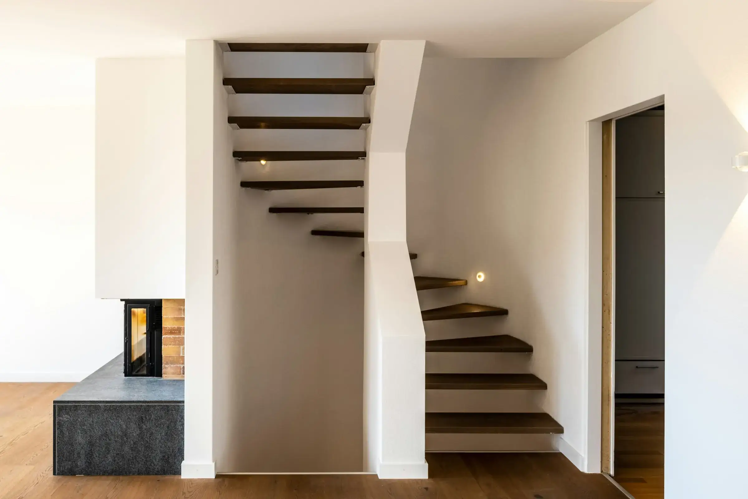 After: open main floor with oak flooring, a floating staircase with dark oak treads and step lights, and a stone-clad fireplace.