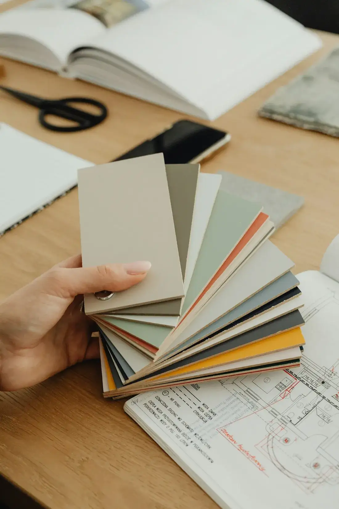 A hand fanning out paint and material swatches over a floor plan and an open notebook.