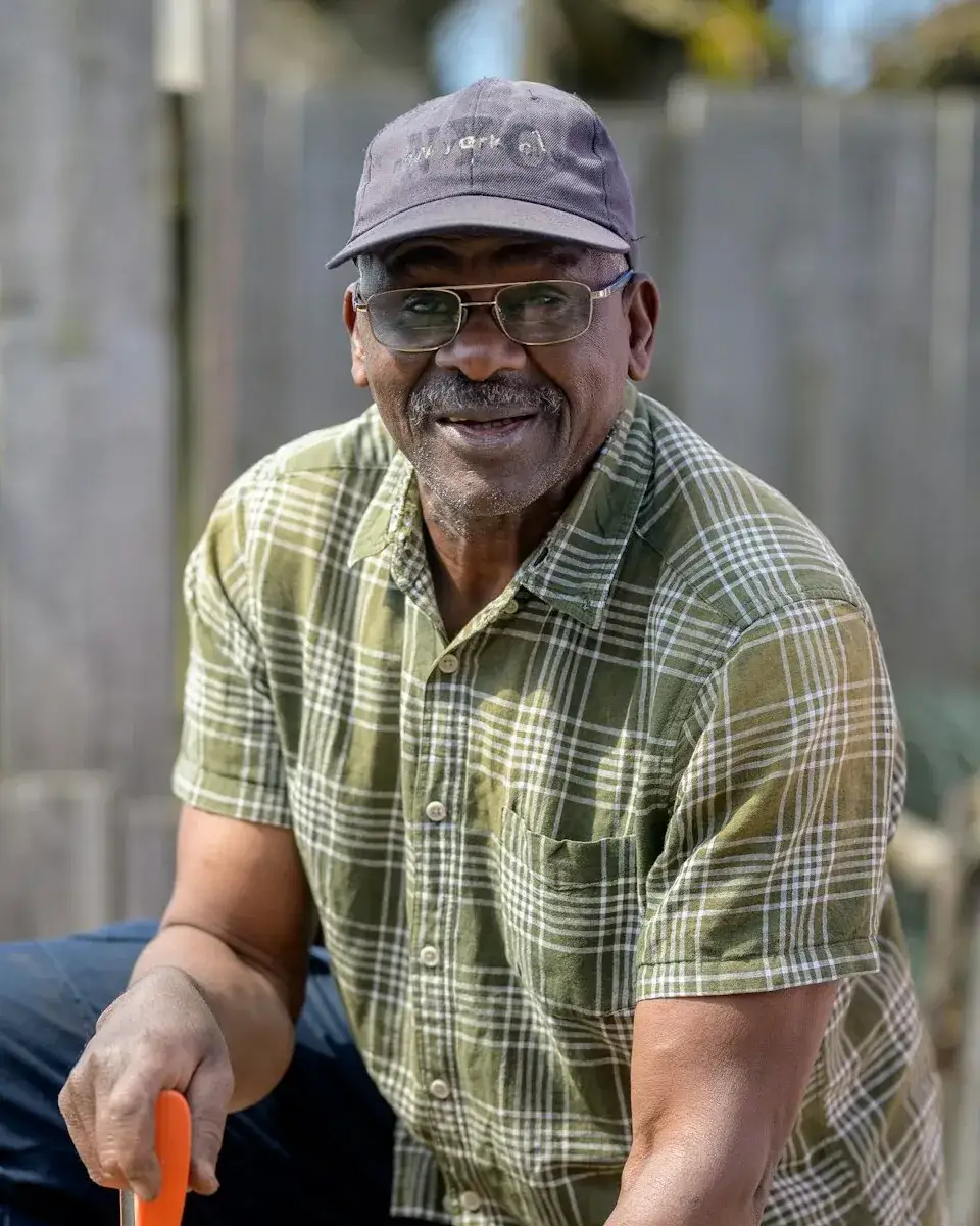 Winston Clarke, smiling in a cap and plaid shirt, with a hand saw at an outdoor work bench.