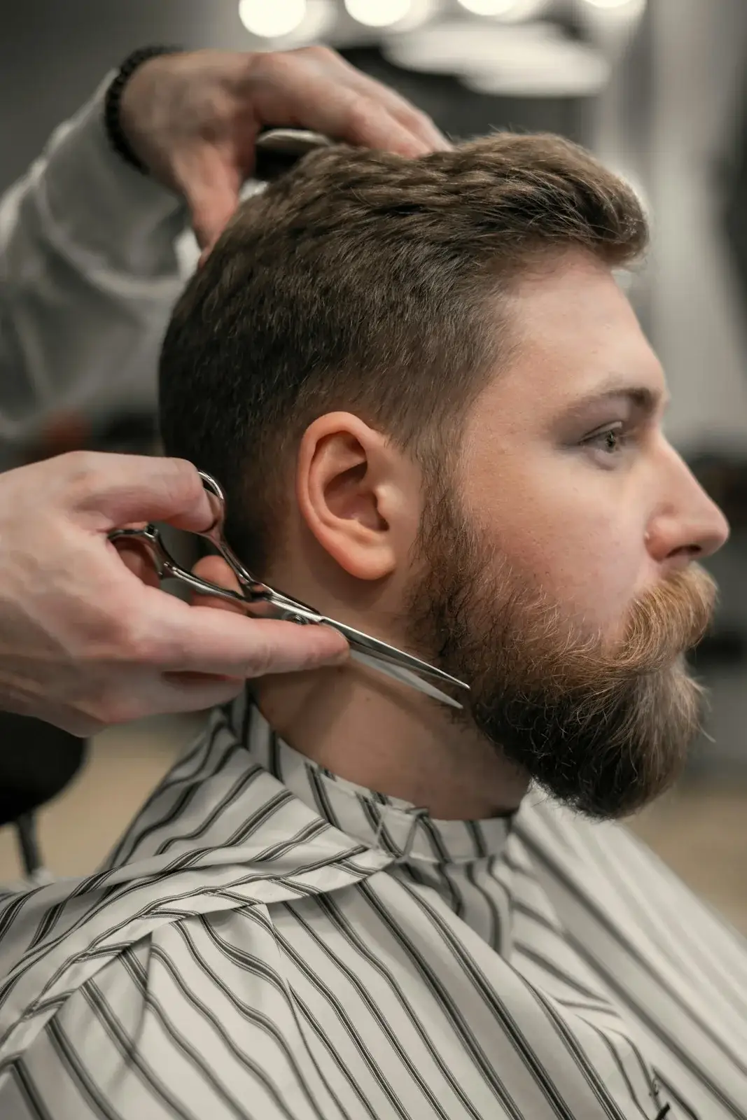 Barber trimming a full beard with scissors while the client wears a striped cape.