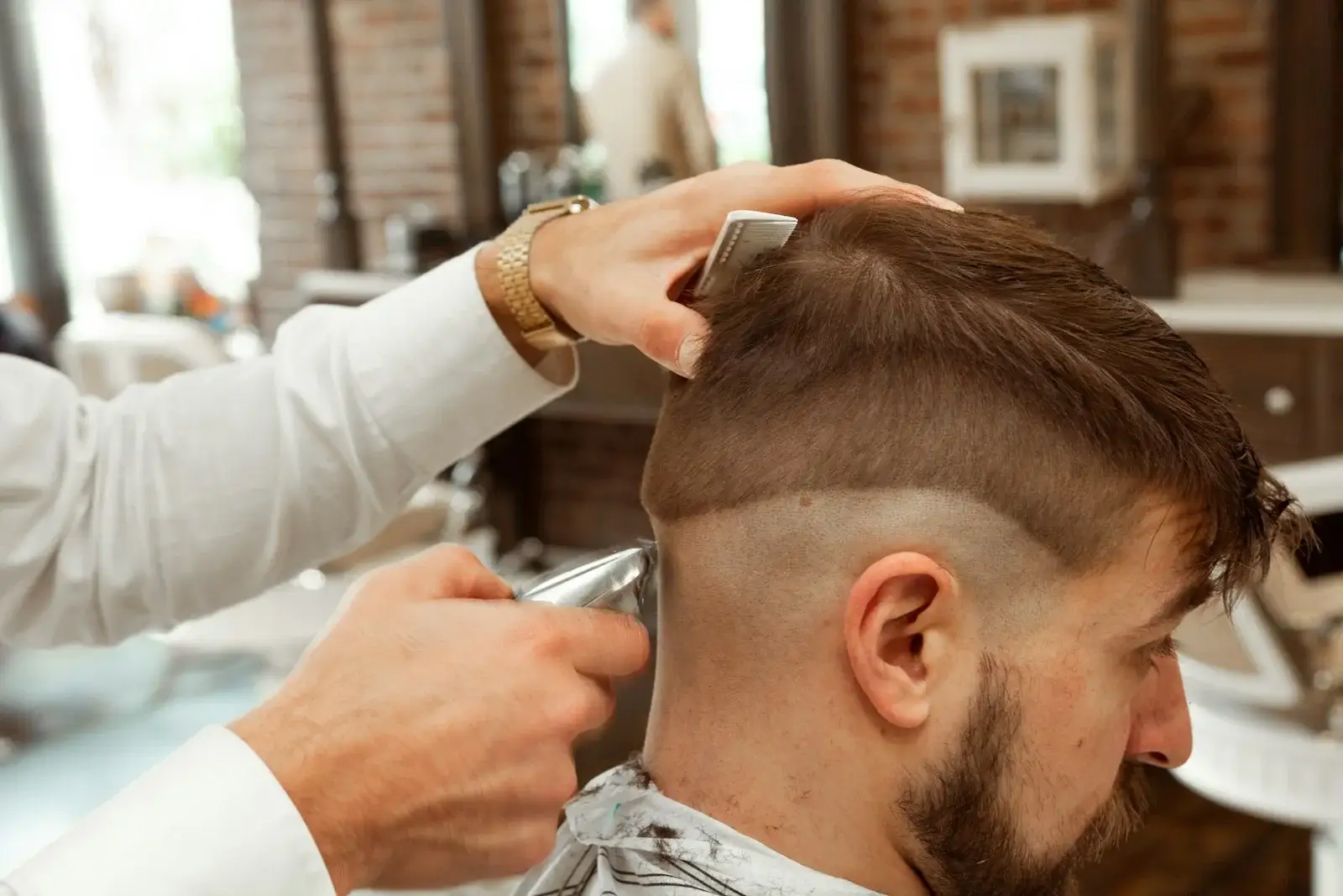 Barber in a white shirt shaping a skin fade with clippers on a bearded client.
